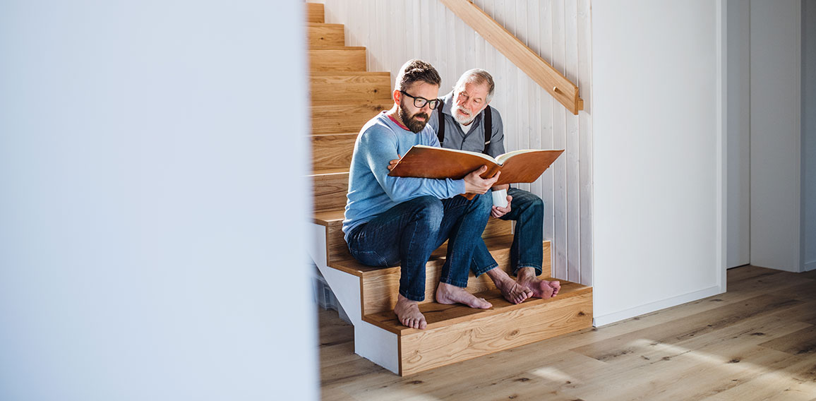 An adult son and senior father sitting on stairs indoors at home, looking at photographs.