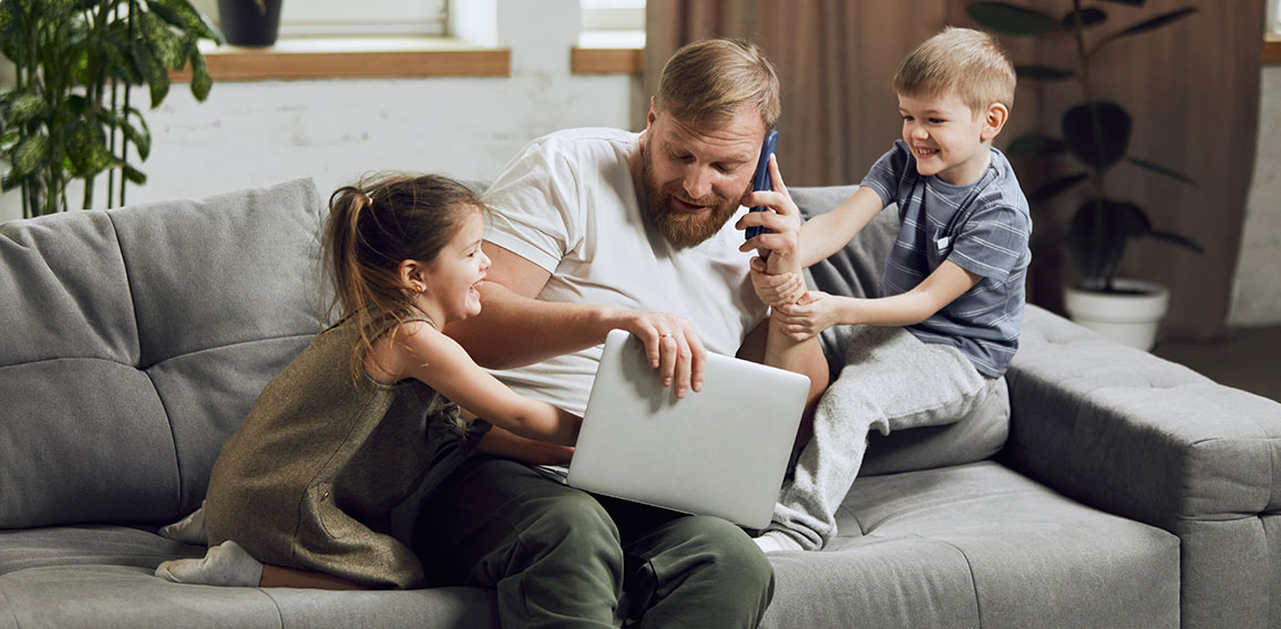 Man, father sitting on couch, talking on phone and working on laptop in living room. Kids bothering and playing with him