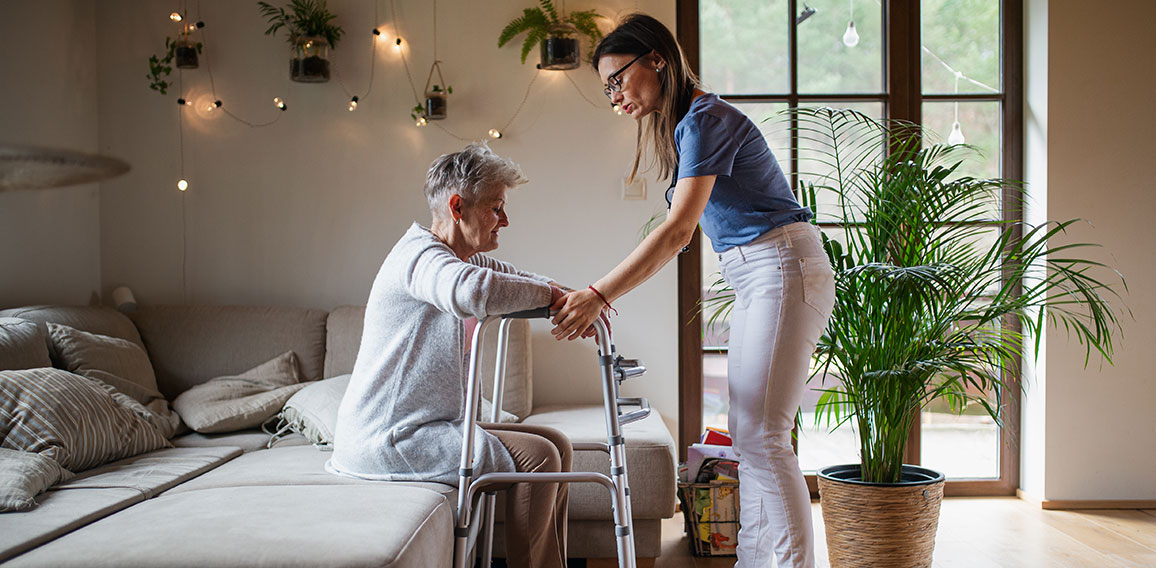 Side view of healthcare worker or caregiver visiting senior woman indoors at home, helping her to walk.