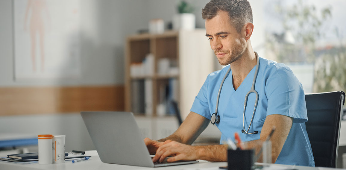 Experienced Male Nurse Wearing Blue Uniform Working on Laptop Co