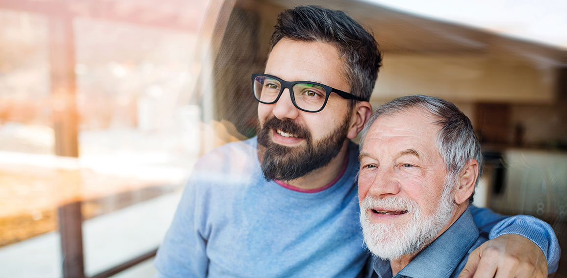 Adult son and senior father indoors at home, looking out. Shot through glass.