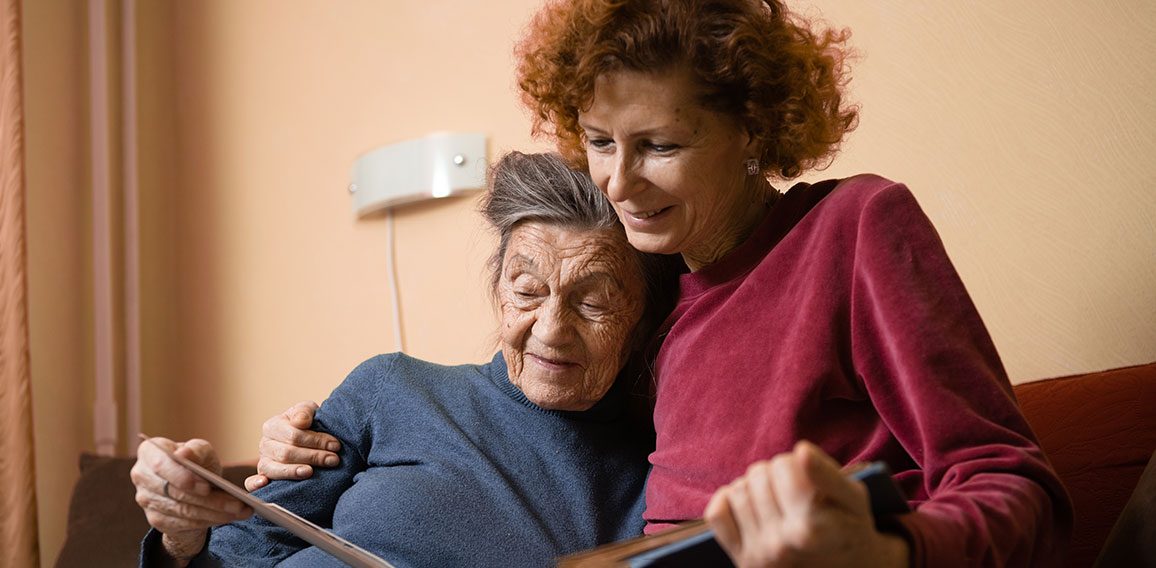 Senior woman and her adult daughter looking at photo album together on couch in living room, talking joyful discussing memories. Weekend with parents, family day, thanksgiving, mom's holiday